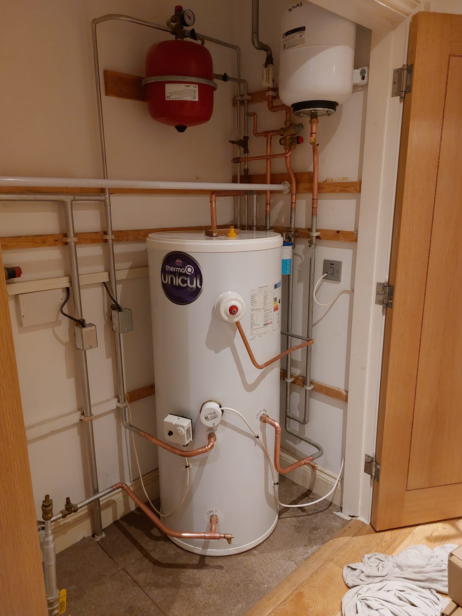 Photograph of a domestic central-heating installation: a white hot-water cylinder branded ‘ThermaQ Unicyl’ sits in a small utility cupboard, surrounded by copper and plastic pipework, valves and fittings, plus a red expansion vessel and a white wall-mounted tank — part of a modern UK home central heating system.